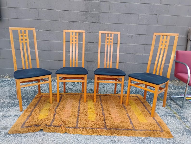 May include: A set of four wooden chairs with high backs featuring a grid pattern at the top and dark blue upholstered seats. The chairs have light brown wooden frames with straight legs and horizontal support bars between the legs for stability.