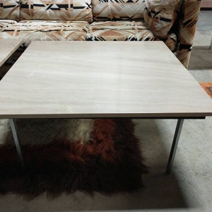 May include: A light-colored, rectangular coffee table with a smooth surface and metal legs. The table is set on a shaggy, brown and red rug. The table's design is simple and modern.