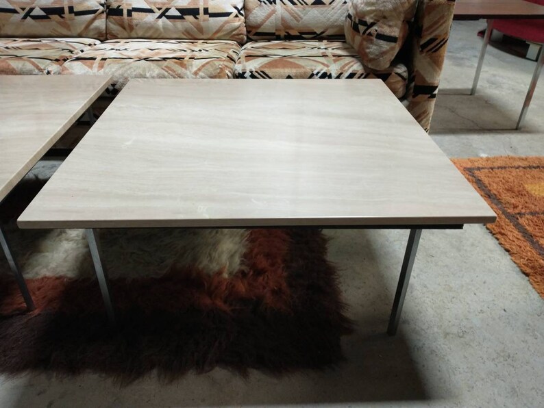 May include: A light-colored, rectangular coffee table with a smooth surface and metal legs. The table is set on a shaggy, brown and red rug. The table's design is simple and modern.