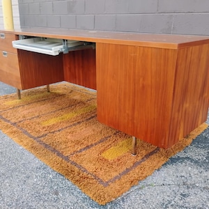 May include: A long wooden desk with a smooth brown finish, featuring a set of drawers on the left side and a sliding white keyboard tray mounted underneath the desktop. The desk is supported by thin, metallic legs and has a rectangular shape.