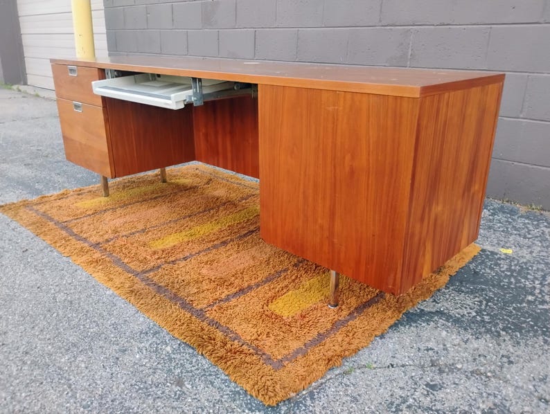 May include: A long wooden desk with a smooth brown finish, featuring a set of drawers on the left side and a sliding white keyboard tray mounted underneath the desktop. The desk is supported by thin, metallic legs and has a rectangular shape.