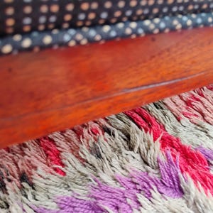 K&ouml;nnte beinhalten: Ein polierter rotbrauner Holzrahmen liegt &uuml;ber einem strukturierten Teppich mit Hochflor in Creme-, Rosa- und Violettt&ouml;nen. &Uuml;ber dem Holzrahmen zeigt ein dunkles Stoffkissen ein sich wiederholendes Muster aus kleinen wei&szlig;en Punkten.