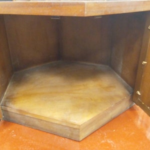 May include: A wooden corner cabinet with two open doors. The interior is a light brown color, with a hexagonal base. The cabinet is made of wood and has a warm, natural finish. The doors are open, revealing the empty storage space.