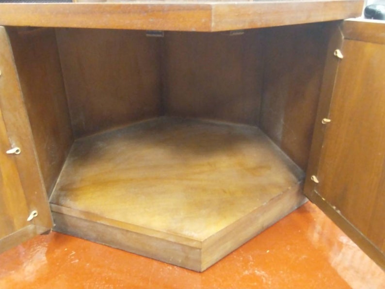 May include: A wooden corner cabinet with two open doors. The interior is a light brown color, with a hexagonal base. The cabinet is made of wood and has a warm, natural finish. The doors are open, revealing the empty storage space.