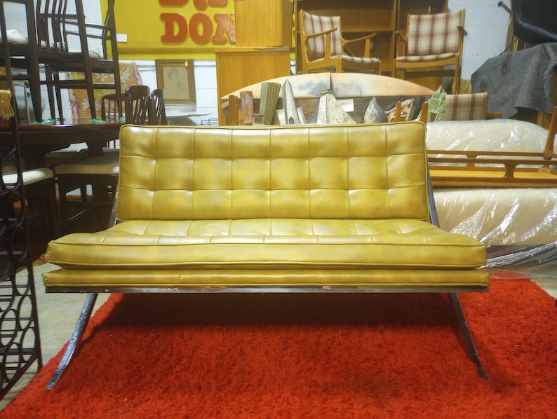 K&ouml;nnte beinhalten: Ein Vintage-Sofa aus gelbem Leder mit einem gesteppten Design und einem Metallrahmen. Das Sofa steht auf einem roten Teppich.