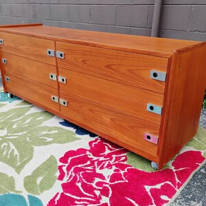 May include: A long, rectangular wooden dresser with a warm brown wood grain finish. It features six drawers arranged in two vertical columns, each equipped with rectangular silver metal pulls. The unit is supported by small wheels at the base.