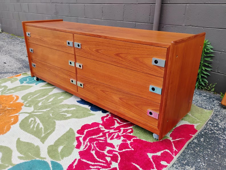 May include: A long, rectangular wooden dresser with a warm brown wood grain finish. It features six drawers arranged in two vertical columns, each equipped with rectangular silver metal pulls. The unit is supported by small wheels at the base.