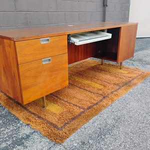 May include: A rectangular wooden desk with a warm brown wood grain finish. The desk features two stacked drawers on the left side with silver metal pulls, a central sliding keyboard tray, and a cabinet on the right side, all supported by thin legs.