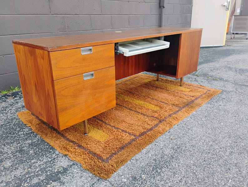May include: A rectangular wooden desk with a warm brown wood grain finish. The desk features two stacked drawers on the left side with silver metal pulls, a central sliding keyboard tray, and a cabinet on the right side, all supported by thin legs.