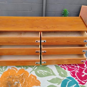 May include: A rectangular wooden dresser with a light brown finish featuring six open drawers arranged in two vertical columns. Each drawer is equipped with a rectangular silver metal pull handle on the left and right sides of the drawer front.