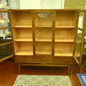 May include: A wooden cabinet with glass doors and three drawers. The cabinet has a light wood finish and is empty.