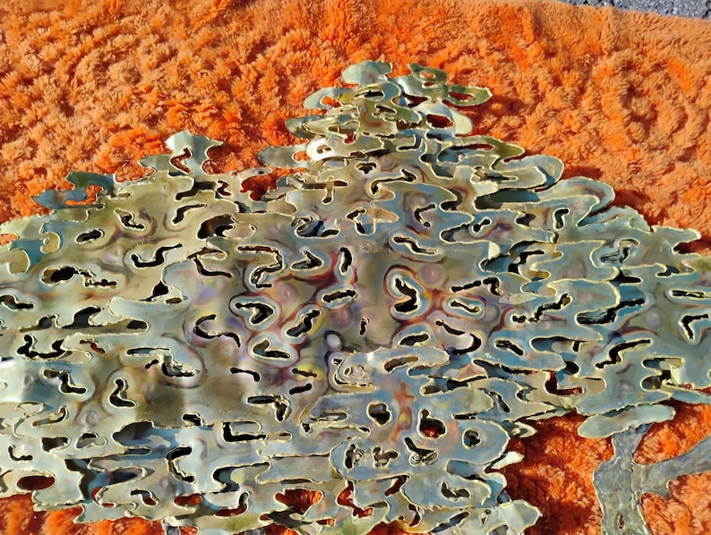 May include: A large, abstract wall sculpture made of layered, cut metal. The metal has a gold finish with iridescent hues and a textured, organic pattern.