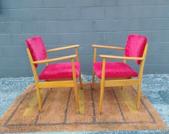 Könnte beinhalten: Zwei Mid-Century-Modern-Sessel mit hellen Holzrahmen und leuchtend roter Samtpolsterung. Die Stühle stehen auf einem braunen Teppich mit geometrischem Muster. Die Stühle haben Armlehnen und stehen vor einer grauen Backsteinmauer.