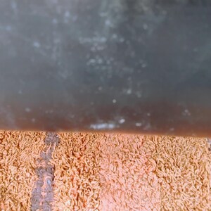 May include: A rectangular bath mat featuring a shaggy, textured pile in a striped pattern of muted orange, peach, and dark brown tones. The surface consists of dense, looped fibers that create a soft, uneven appearance across the entire mat.