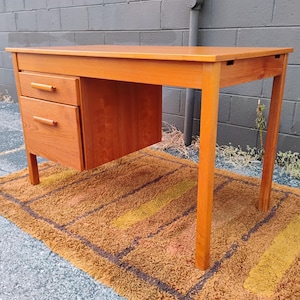 Op de afbeelding: Een lichtbruin houten bureau met een rechthoekig blad en een ladeblok aan de linkerkant. Het ladeblok bevat een kleinere bovenste lade en een grotere onderste lade, beide voorzien van horizontale houten handgrepen en rechte poten.