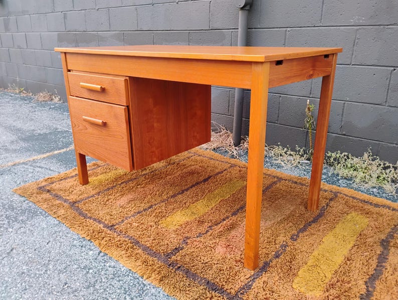Op de afbeelding: Een lichtbruin houten bureau met een rechthoekig blad en een ladeblok aan de linkerkant. Het ladeblok bevat een kleinere bovenste lade en een grotere onderste lade, beide voorzien van horizontale houten handgrepen en rechte poten.