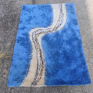 Puede incluir: Una alfombra rectangular de pelo largo con un color base azul vibrante y un patr&oacute;n ondulado prominente en crema y marr&oacute;n oscuro que recorre verticalmente el centro. La alfombra tiene una superficie de pelo grueso y texturizado.