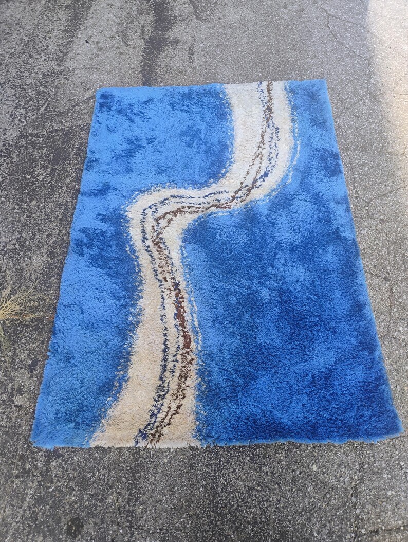 Puede incluir: Una alfombra rectangular de pelo largo con un color base azul vibrante y un patr&oacute;n ondulado prominente en crema y marr&oacute;n oscuro que recorre verticalmente el centro. La alfombra tiene una superficie de pelo grueso y texturizado.