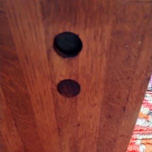 May include: Close-up of a wooden surface with two circular holes. The wood grain is visible, and the holes are dark, contrasting with the lighter wood. The image focuses on the texture and detail of the wood.