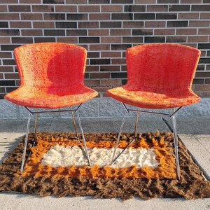 May include: Two orange mid-century modern chairs with chrome legs are displayed on a brown, orange, and white rug. The chairs have a curved back and seat. The rug has a rectangular shape and a shaggy texture. The chairs are placed in front of a brick wall.
