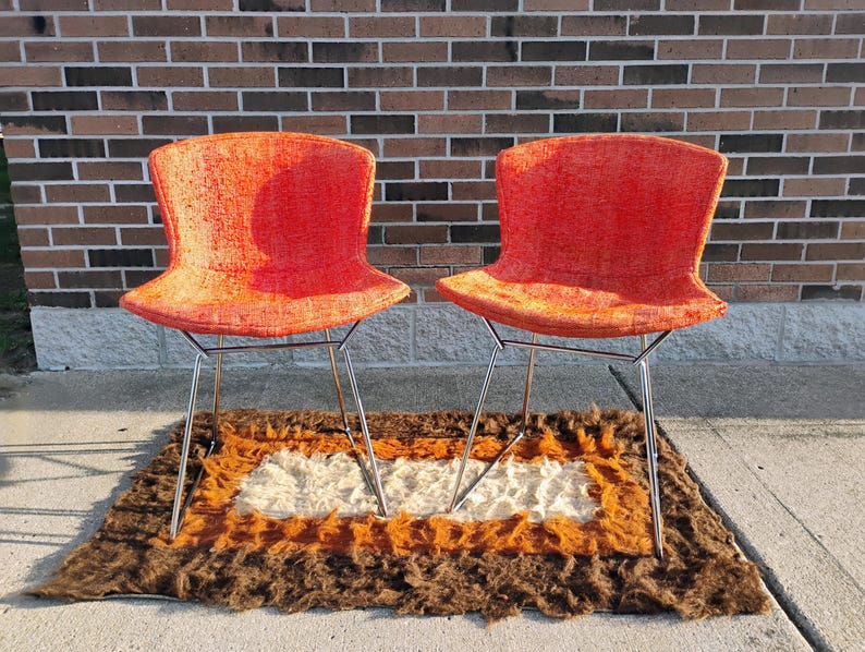 May include: Two orange mid-century modern chairs with chrome legs are displayed on a brown, orange, and white rug. The chairs have a curved back and seat. The rug has a rectangular shape and a shaggy texture. The chairs are placed in front of a brick wall.
