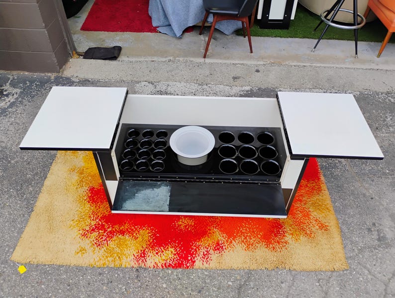 May include: A black and white bar cart with two fold-down serving trays and a built-in ice bucket. The cart has a black and white checkered pattern on the bottom.