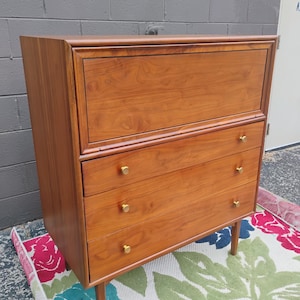 May include: A mid-century modern wooden dresser with three drawers and a top compartment. The dresser is a warm brown color with gold-colored knobs and sits on four tapered legs. It is placed on a floral patterned rug.