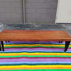K&ouml;nnte beinhalten: Ein Mid-Century-Modern-Bank mit einer Lattenoberfl&auml;che aus Holz und schwarzen Beinen. Die Bank steht auf einem bunten gestreiften Teppich mit gelben, gr&uuml;nen, violetten und roten B&auml;ndern. Die Bank ist etwa 152 cm lang.