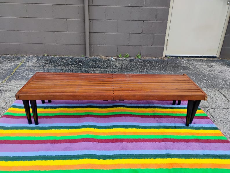 K&ouml;nnte beinhalten: Ein Mid-Century-Modern-Bank mit einer Lattenoberfl&auml;che aus Holz und schwarzen Beinen. Die Bank steht auf einem bunten gestreiften Teppich mit gelben, gr&uuml;nen, violetten und roten B&auml;ndern. Die Bank ist etwa 152 cm lang.