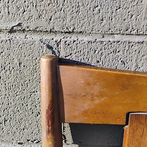 May include: Close-up of the top corner of a wooden chair with a light brown finish. The chair has a simple design with straight lines and a smooth surface.