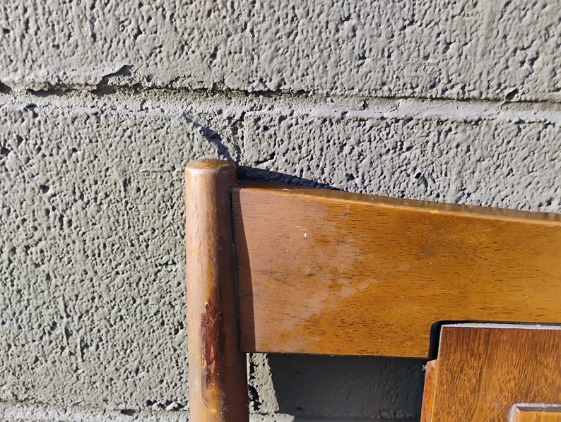 May include: Close-up of the top corner of a wooden chair with a light brown finish. The chair has a simple design with straight lines and a smooth surface.