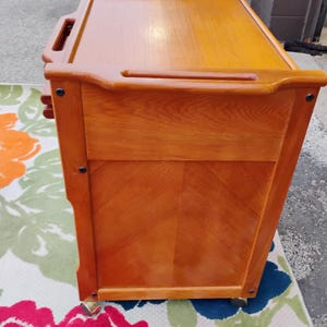 May include: A wooden serving cart with three drawers and wheels. The cart is made of light brown wood and has a dark brown top. The cart is on a floral rug.