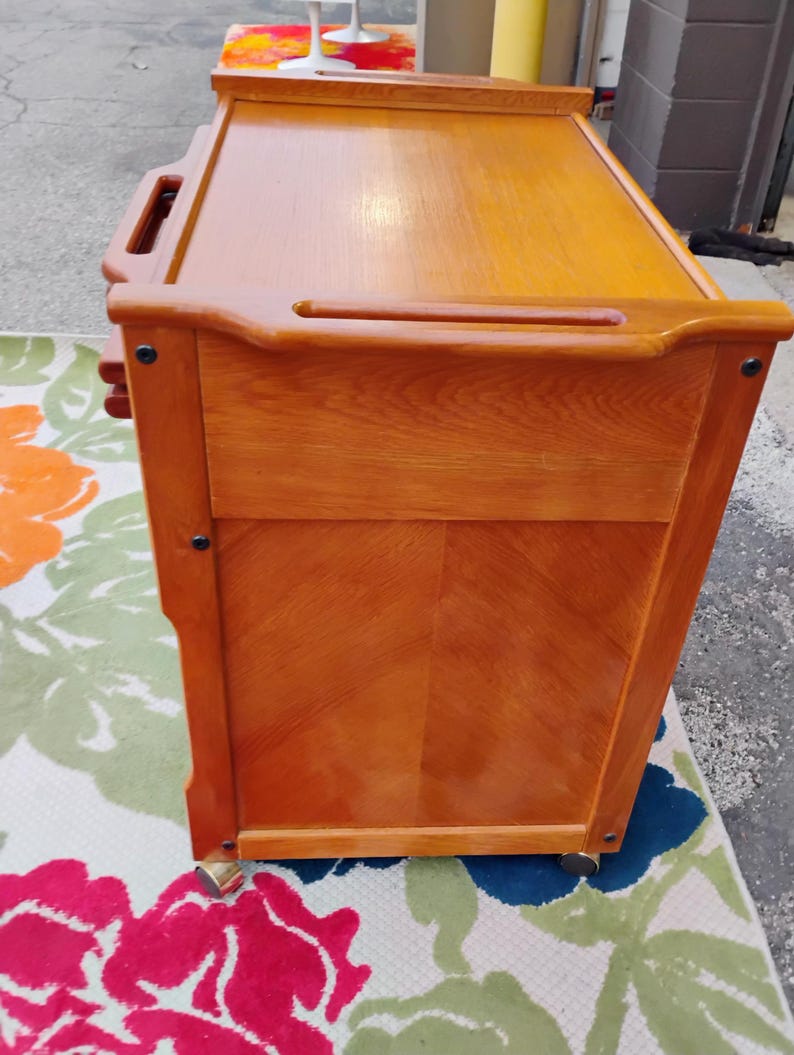 May include: A wooden serving cart with three drawers and wheels. The cart is made of light brown wood and has a dark brown top. The cart is on a floral rug.