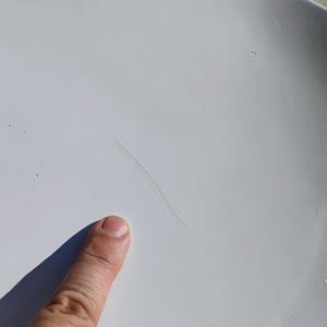 K&ouml;nnte beinhalten: Nahaufnahme einer wei&szlig;en Oberfl&auml;che mit einer langen, d&uuml;nnen, dunklen, haar&auml;hnlichen Markierung. Ein Finger zeigt auf die Oberfl&auml;che und wirft einen Schatten. Die Oberfl&auml;che erscheint glatt und leicht gekr&uuml;mmt.