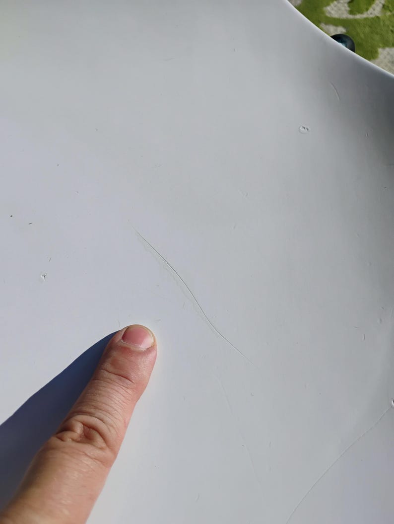 K&ouml;nnte beinhalten: Nahaufnahme einer wei&szlig;en Oberfl&auml;che mit einer langen, d&uuml;nnen, dunklen, haar&auml;hnlichen Markierung. Ein Finger zeigt auf die Oberfl&auml;che und wirft einen Schatten. Die Oberfl&auml;che erscheint glatt und leicht gekr&uuml;mmt.