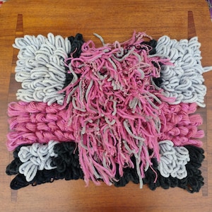 May include: A handmade textile art piece with a rectangular design. The piece features a combination of pink, gray, and black yarn, creating a textured, abstract pattern. The artwork is placed on a wooden surface.