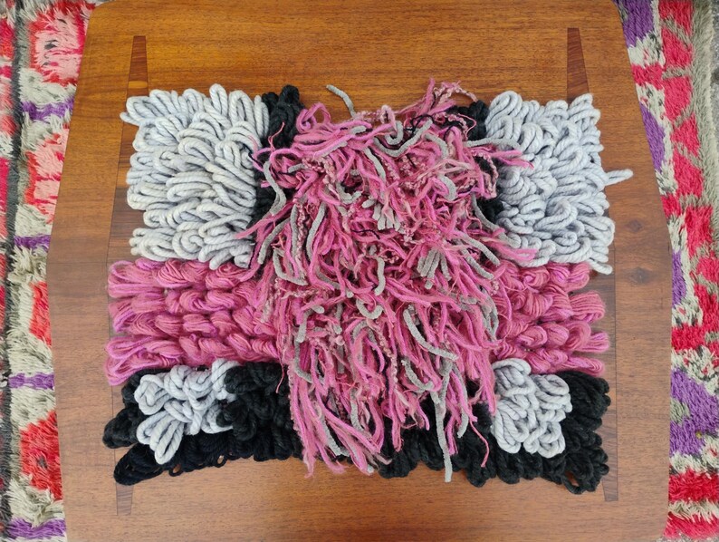 May include: A handmade textile art piece with a rectangular design. The piece features a combination of pink, gray, and black yarn, creating a textured, abstract pattern. The artwork is placed on a wooden surface.