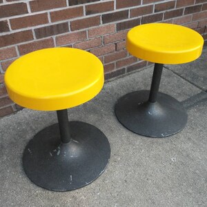 May include: Two yellow round stools with black bases. The stools are made of plastic and metal.