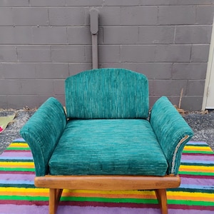 May include: An armchair upholstered in a textured teal green fabric with vertical white striations. The chair features a curved backrest, wide armrests, and a wooden frame with four legs. The fabric shows visible wear along the right armrest edge.