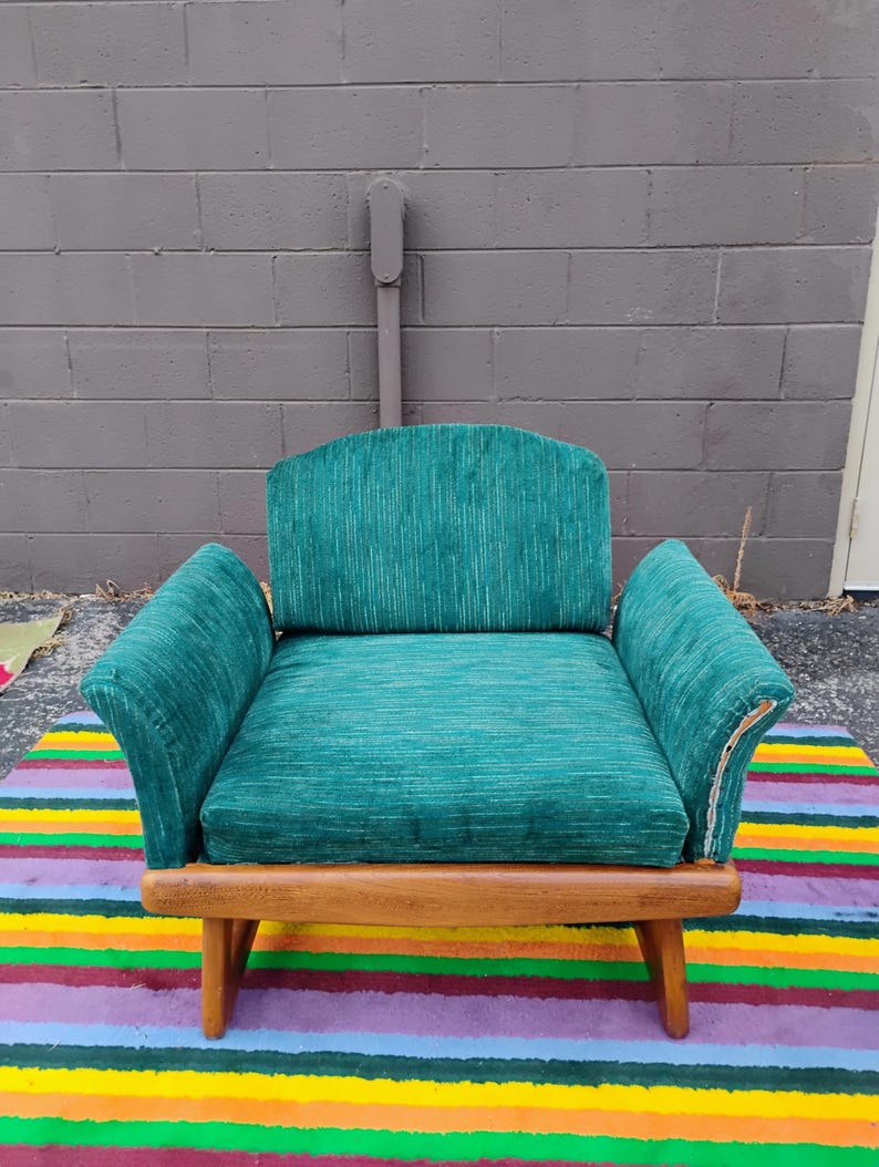 May include: An armchair upholstered in a textured teal green fabric with vertical white striations. The chair features a curved backrest, wide armrests, and a wooden frame with four legs. The fabric shows visible wear along the right armrest edge.