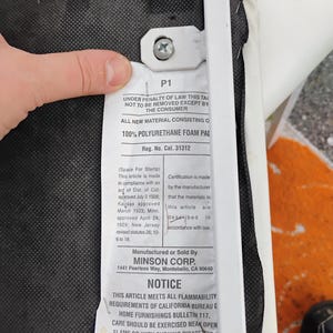 May include: A white tag attached to a chair frame, with text including "100% POLYURETHANE FOAM PAD" and "NOTICE: THIS ARTICLE MEETS ALL FLAMMABILITY REQUIREMENTS OF CALIFORNIA BUREAU". The tag is held by a hand.