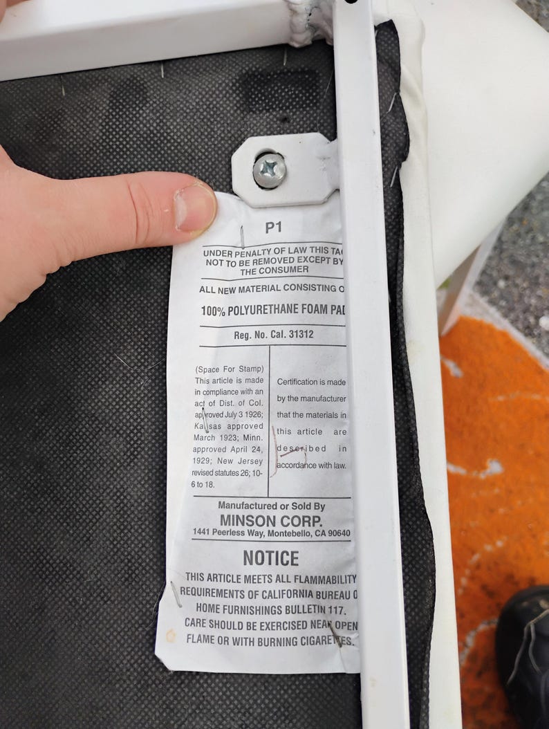 May include: A white tag attached to a chair frame, with text including "100% POLYURETHANE FOAM PAD" and "NOTICE: THIS ARTICLE MEETS ALL FLAMMABILITY REQUIREMENTS OF CALIFORNIA BUREAU". The tag is held by a hand.