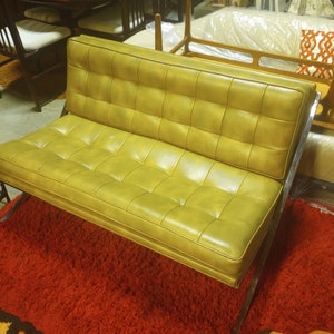 K&ouml;nnte beinhalten: Ein Vintage-Zweisitzer-Sofa aus gr&uuml;nem Leder mit Steppdesign und verchromten Beinen. Das Sofa steht auf einem roten Shaggy-Teppich.