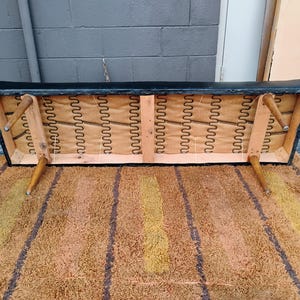May include: The underside of a black rectangular bench showing a wooden frame, four tapered light brown wooden legs, and exposed metal sinuous springs attached to a tan fabric backing. The structure is visible from a low angle view.