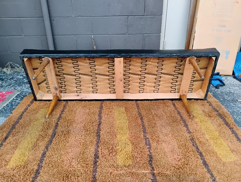 May include: The underside of a black rectangular bench showing a wooden frame, four tapered light brown wooden legs, and exposed metal sinuous springs attached to a tan fabric backing. The structure is visible from a low angle view.