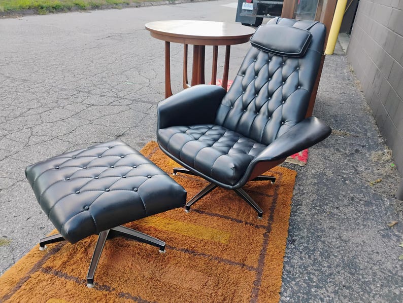 May include: A black tufted lounge chair with a matching ottoman, both featuring button detailing and a headrest. The chair and ottoman are supported by silver-toned metal swivel bases with four legs, providing a stable foundation for the seating.