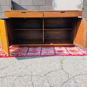 Op de afbeelding: Een rechthoekig houten dressoir met twee bovenste lades met verzonken grepen en twee zijkasten met draaideuren. Het interieur bevat een centrale plank die de opbergruimte verdeelt. Het hout heeft een warme bruine afwerking met nerven.