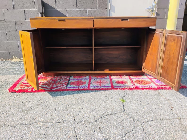 Op de afbeelding: Een rechthoekig houten dressoir met twee bovenste lades met verzonken grepen en twee zijkasten met draaideuren. Het interieur bevat een centrale plank die de opbergruimte verdeelt. Het hout heeft een warme bruine afwerking met nerven.
