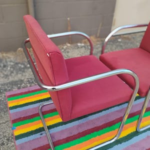 May include: Two armchairs featuring deep red fabric upholstery on the seat and backrest, supported by a continuous, polished chrome tubular metal frame that forms a cantilevered base and integrated armrests for a sleek, structural appearance.