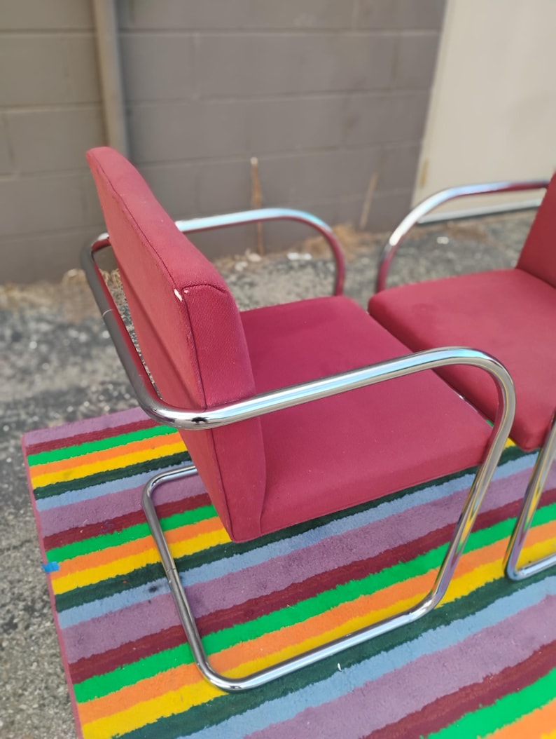 May include: Two armchairs featuring deep red fabric upholstery on the seat and backrest, supported by a continuous, polished chrome tubular metal frame that forms a cantilevered base and integrated armrests for a sleek, structural appearance.